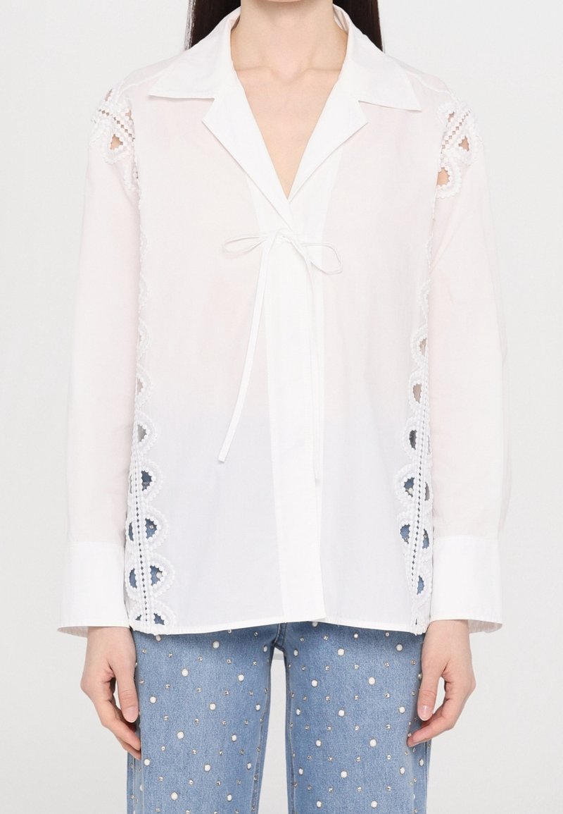 Femme portant un chemisier blanc à manches longues avec bordure en dentelle et nœud à l'avant, associé à un jean bleu orné de perles blanches.