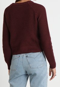 Bordowy sweter dziany z teksturowanym wzorem, z prążkowanym dołem i mankietami, noszony z jasnoniebieskimi dżinsami. Tył ozdobiony jest rękawami raglan.