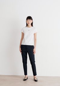 White cotton t-shirt with small blue logo, paired with slim-fit black pants. Model wears black flats, with straight dark hair and minimal accessories.