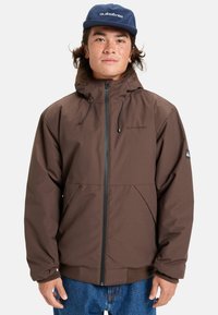 Ung man med långt hår iklädd en brun jacka med huva och en blå Quiksilver-keps, stående mot en vit bakgrund.