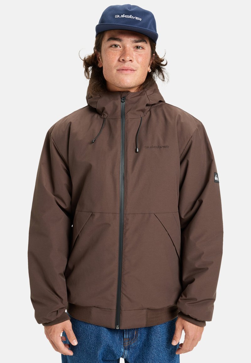 Ung man med långt hår iklädd en brun jacka med huva och en blå Quiksilver-keps, stående mot en vit bakgrund.