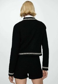 Czarna krótka kurtka z teksturowanej tkaniny z srebrnym wykończeniem przy kołnierzu i dole, zestawiona z dopasowanymi czarnymi szortami.