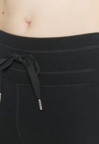 Černé elastické kalhoty v pase se stahovací šňůrkou. Mají kontrastní švy a malé značkové štítky. Měkký, pružný materiál s hladkou texturou.