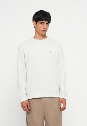 Lichtgrijs sweatshirt met ronde hals, lange mouwen en een subtiel logo op de borst. De stof heeft een gladde textuur, gecombineerd met beige broek.
