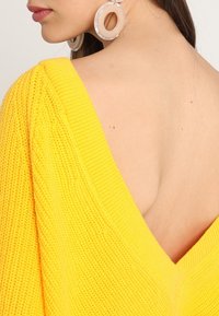 Żółty sweter na drutach z głębokim dekoltem w kształcie litery V. Tkanina ma teksturowany wzór. Wygląd uzupełniają przezroczyste, okrągłe kolczyki.