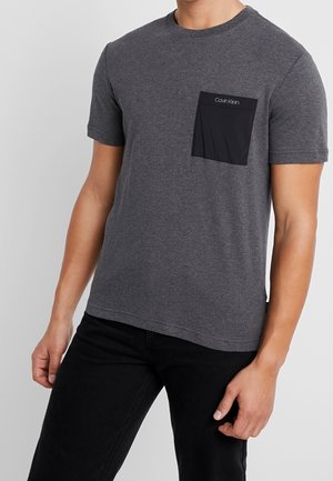 Šedé bavlněné tričko s kulatým výstřihem a krátkým rukávem, má černou kapsu s logem. Tkanina má hladkou strukturu.