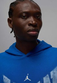 Niebieska bluza z kapturem z nadrukiem na przodzie. Posiada dekolt w kształcie litery V, sznurek do ściągania kaptura oraz biały logo Air Jordan na piersi.