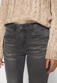 Jeans de mezclilla gris con un diseño ajustado, que cuentan con bolsillos delanteros y cierre de botón metálico, combinados con un suéter de punto beige texturizado.