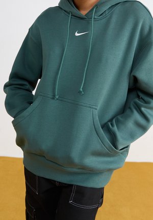 Person iført teal Nike-hoodie med forlomme og snoretræk, kombineret med sorte bukser med hvide syninger.