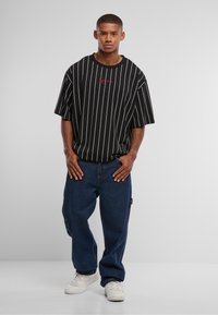 Junger Mann steht mit den Händen in der Nähe der Tasche, trägt ein schwarzes, gestreiftes, übergroßes T-Shirt, lockere blaue Jeans und weiße Turnschuhe vor einem schlichten grauen Hintergrund.