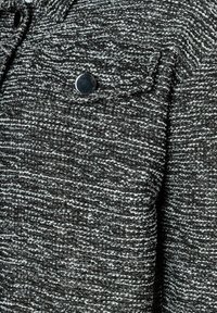 Schwarz-weißer strukturierter Stoff mit einem gewebten Muster, das einen auffälligen Knopf und ein einzelnes Brusttaschendetail aufweist.