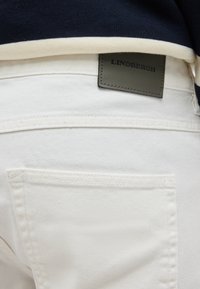 Τσέπη πίσω λευκού τζιν με μαύρη ετικέτα μάρκας Lindbergh, φορεμένη με σκουρόχρωμο μπλε τοπ που έχει λευκή ρίγα στο τελείωμα.