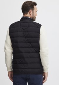 Svart puffer vest med quiltet design, høy krage og uten ermer, båret over en kremfarget genser. Synlig søm og teksturert stoff.