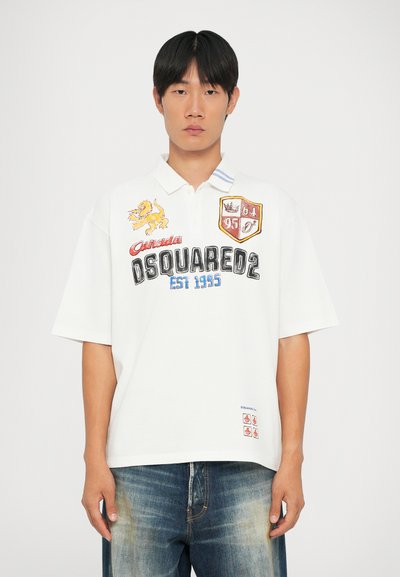 Cămașă polo albă cu logo-uri brodate, inclusiv un leu, un blazon cu numerele 64 și 95 și un etichetă pe mânecă. Se potrivește perfect cu blugi din denim.