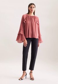 Blusa plissettata color lampone con dettagli arricciati, maniche lunghe a campana; abbinata a pantaloni aderenti neri e tacchi neri con cinturini.