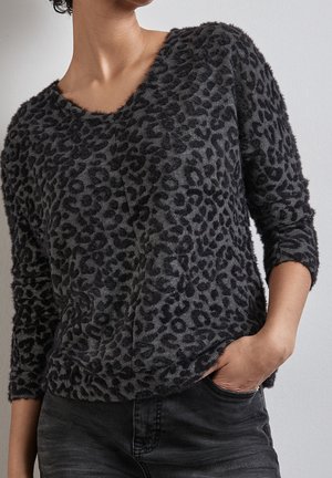 Vrouw draagt een donzig donkergrijs trui met luipaardprint, een ronde hals, lange mouwen en zwarte spijkerbroek, hand in de zak.