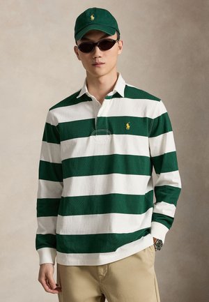 Uomo che indossa una polo a maniche lunghe a righe verdi e bianche, berretto verde, occhiali da sole scuri, pantaloni beige e un orologio da polso su uno sfondo semplice.