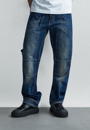 Jean en denim bleu foncé avec poches avant inclinées, détails en patch aux genoux, baskets noires, et un bas de chemise blanc.