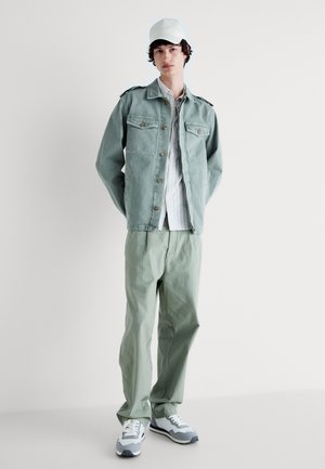 Chaqueta de denim verde clara con cierre de botones y dos bolsillos en el pecho, combinada con una camisa a rayas y pantalones sueltos de color verde claro. Gorra blanca y zapatillas.