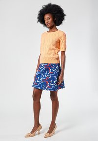 Vaalean oranssi lyhythihainen neulepaita, jossa on tekstuuri, yhdistettynä siniseen hameeseen, jossa on värikkäitä abstrakteja muotoja, sekä nude korkeakorkoiset kengät.