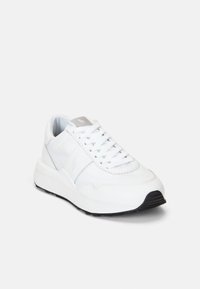 Baskets blanches avec une tige en cuir lisse et textile, comportant un bout rond, des lacets et une semelle en caoutchouc noir texturé. Marquage subtil sur la languette.