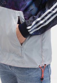 Ruka v zipové kapse světle šedé bundy s nastavitelnou šňůrkou; má na sobě tmavou bundu s bílými pruhy a modré džíny.