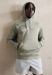 Olivově zelená mikina s kapucí a bílým logem Nike, s klokaní kapsou a stahovací kapucí. Model má na sobě světle šedé šortky se žlutým lemováním.