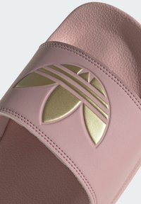 Różowe klapki wykonane z teksturowanego materiału syntetycznego z błyszczącym złotym logo oraz wzorem liści i pasków na górnym pasku.