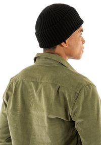 Gorro de punto negro con canalé, combinado con una camisa de pana verde oliva claro, que cuenta con un cuello y rayas verticales en la tela.