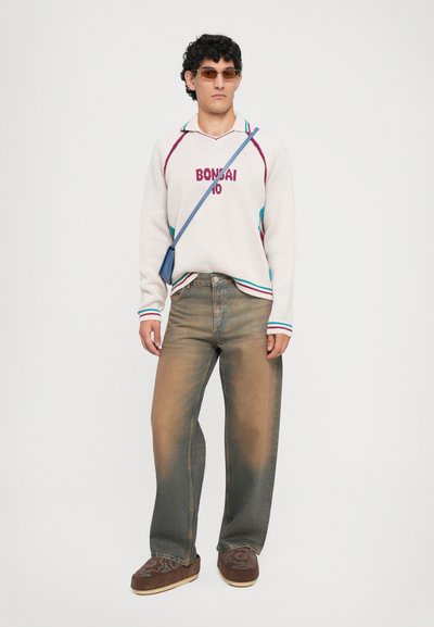 Suéter de punto blanco con "BONSAI 10" en rojo y detalles en azul. Jeans anchos de color marrón desvanecido. Zapatos marrones bordados, gafas de sol ahumadas y bolso cruzado.