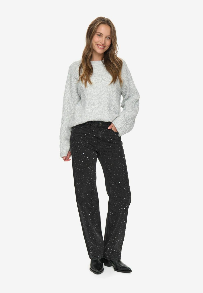 Lächelnde Frau mit langen Haaren, die einen hellgrauen Kabelstrickpullover, schwarze mit Nieten besetzte Jeans und schwarze Stiefel mit Absatz trägt, steht mit einer Hand in der Tasche.
