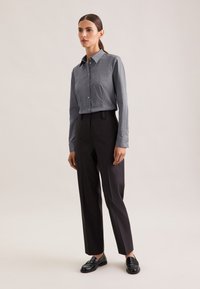 Camicia grigia a maniche lunghe con bottoni e colletto, abbinata a pantaloni neri aderenti e mocassini neri, in piedi su uno sfondo chiaro.