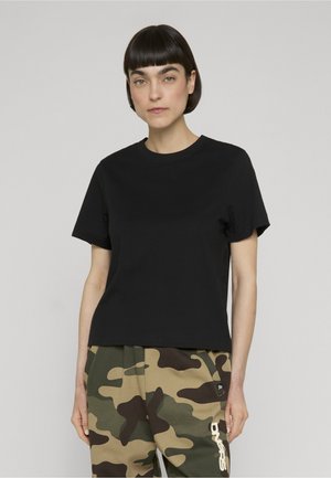 Donna con capelli corti e scuri che indossa una maglietta nera semplice e pantaloni con motivo mimetico, in piedi davanti a uno sfondo chiaro.
