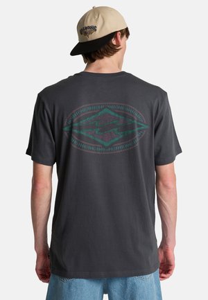 Man draagt een donkergrijs T-shirt met een ovale afbeelding op de achterkant, lichtblauwe spijkerbroek en een beige zwart petje dat achterstevoren is gedragen, gezicht naar achteren.