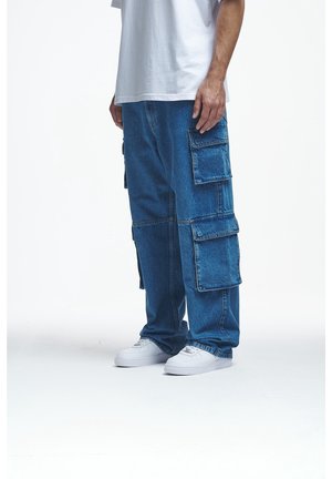 Person trägt lockere blaue Denim-Cargohosen mit mehreren Taschen und weiße Turnschuhe vor weißem Hintergrund.