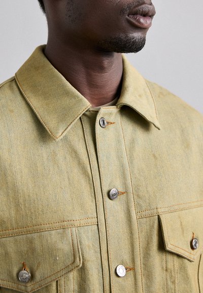 Light green denim shirt with a spread collar, front button closure, and two chest pockets, featuring contrasting orange stitching and silver hardware.
