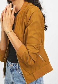 Hnědá bomber bunda s žebrovanými manžetami a lemem, se zipovými bočními kapsami a hladkým, texturovaným materiálem. Nositelná přes černý top.