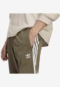 Olivgröna Adidas-byxor med vita tre-randiga detaljer. Mjukt material, elastisk midja, sidorfickor och logotypdetalj på benet.