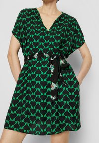 Vestito verde corto con motivo a cuore nero, scollatura a V, maniche corte e una cintura annodata con dettagli floreali. Include tasche laterali.