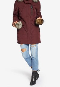 Parka burdeos con detalle de pelo sintético en los puños y la capucha, que cuenta con dos bolsillos laterales y un bolsillo con cremallera, usado sobre unos jeans azul claro desgastados y botas negras.
