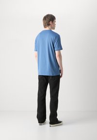Modra bombažna majica s kratkimi rokavi, okroglo izrezana in ohlapnega kroja, v kombinaciji s črnimi jeans hlačami in športnimi copati. Zadnji pogled.