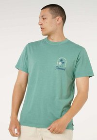 Uomo con t-shirt verde Rip Curl con logo a forma di palma, che guarda a sinistra, indossando pantaloni beige chiari, in piedi su sfondo semplice.