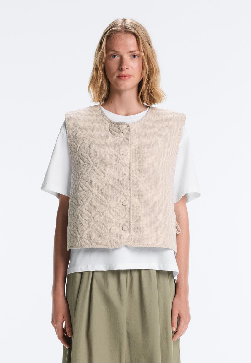 Femme blonde portant une veste matelassée beige sur un t-shirt blanc et un pantalon ample vert olive, debout devant un fond uni.