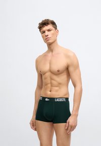 Homme torse nu aux cheveux courts et bouclés portant un boxer Lacoste vert foncé, debout devant un fond clair uni.