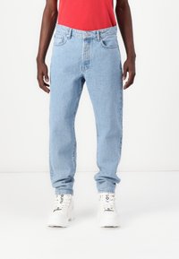 Persona vistiendo jeans azul claro, una camisa roja y zapatillas altas blancas con texto negro en los cordones, de pie contra un fondo blanco.