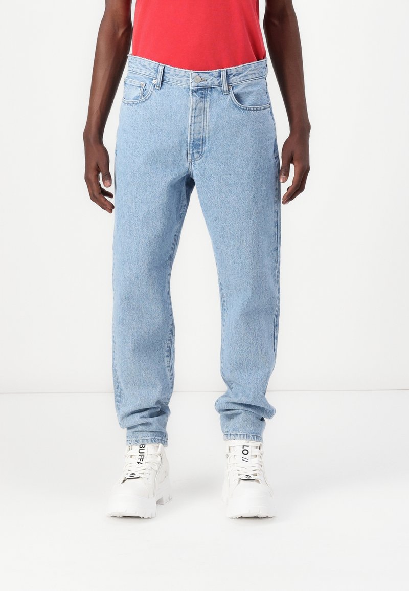 Persona vistiendo jeans azul claro, una camisa roja y zapatillas altas blancas con texto negro en los cordones, de pie contra un fondo blanco.