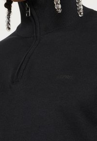 Schwarzer Pullover mit Stehkragen und einem Viertelreißverschluss, aus einem gerippten Strickmaterial gefertigt, mit einem dezenten Logo-Detail auf der Brust.