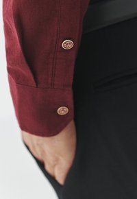 Bordeaux skjorteærme med knapdetaljer i guldmetal. Stoffet ser struktureret ud. Mørkt tøj er synligt i baggrunden.