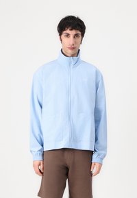 Light blue zip-up jacket with high collar, elastic cuffs, two front pockets, and small logo on the chest. Made from textured fabric.