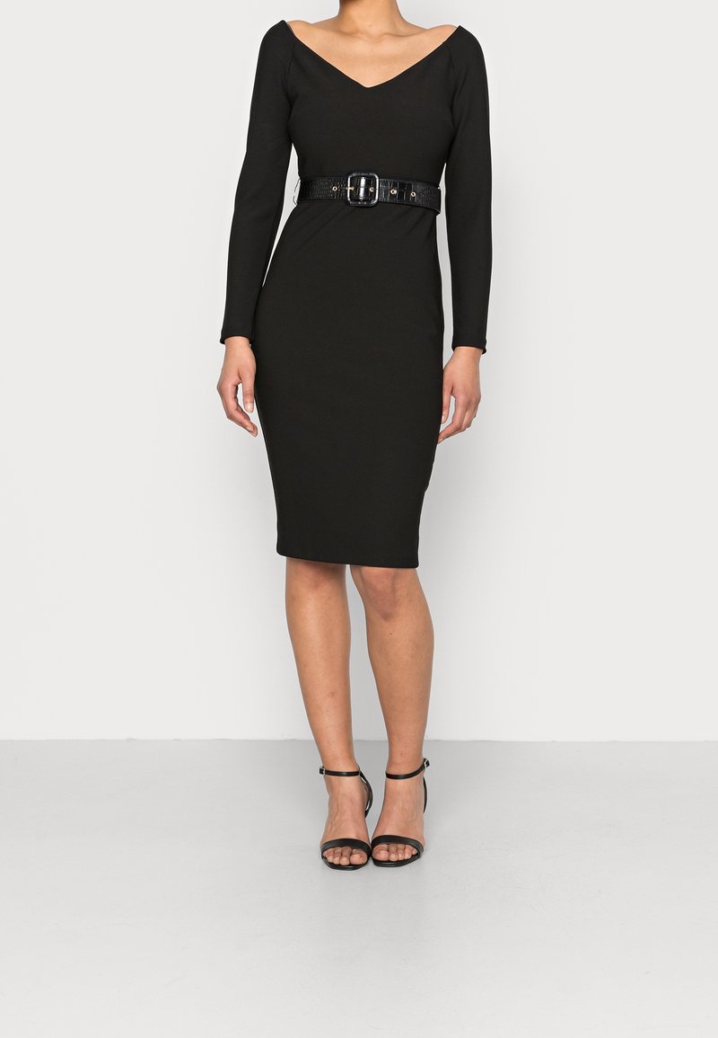 Femme portant une robe noire à manches longues, longueur genoux, avec un col en V, une large ceinture noire et des sandales à talons hauts à bride cheville noires.